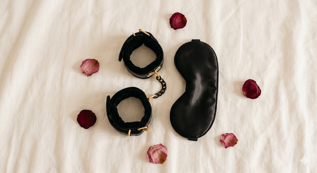 Photographie à plat minimaliste et élégante présentant des menottes doublées de velours noir et un bandeau en satin, disposés sur un tissu ivoire doux avec quelques pétales de roses séchées éparpillés, sous un éclairage chaleureux de style éditorial.